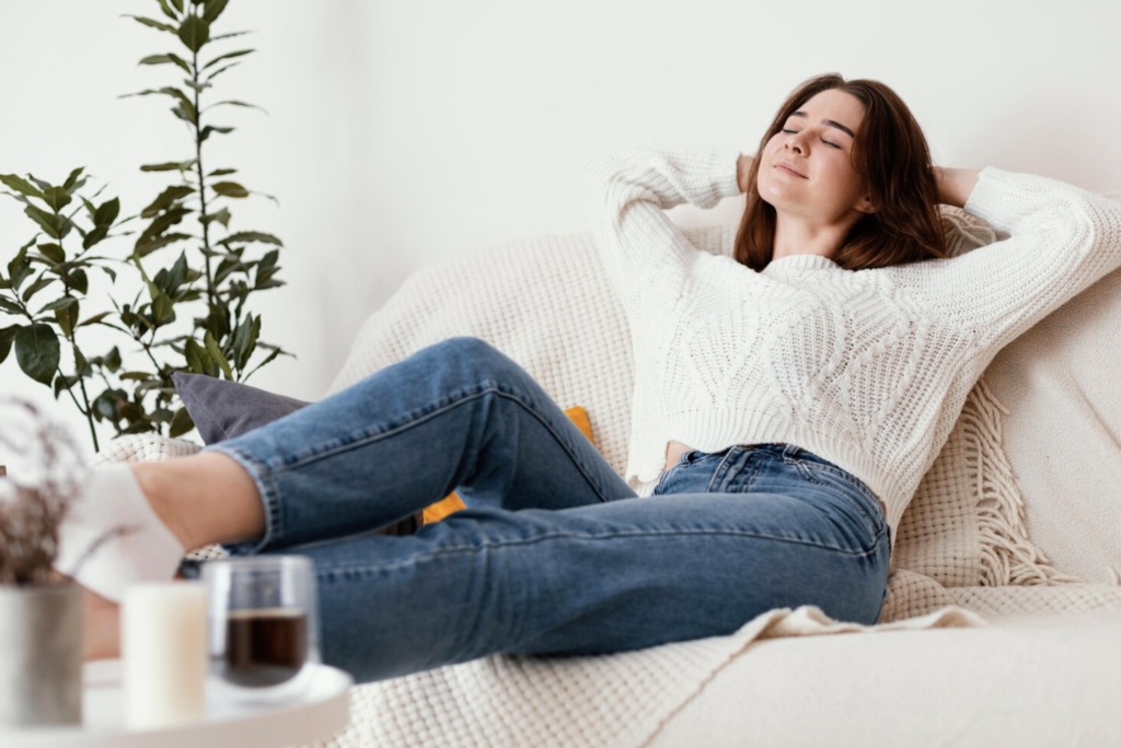 Mujer joven con jersey blanco y jeans relajándose en un sofá beige, disfrutando de un ambiente acogedor con una planta verde en el fondo.